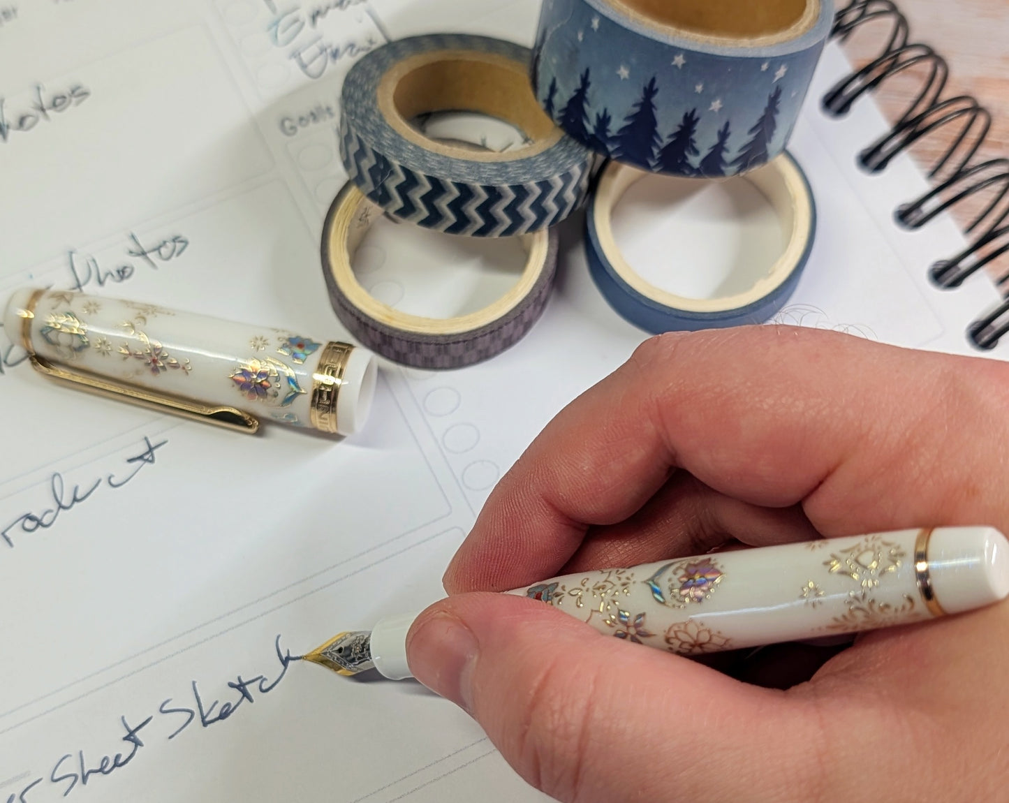 Hand holding a decorative pen writing on paper with washi tape rolls in the background