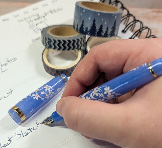Hand holding a purple pen with floral design, surrounded by washi tape rolls on a notebook page.