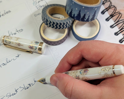 Hand holding a decorative pen writing on paper with washi tape rolls in the background