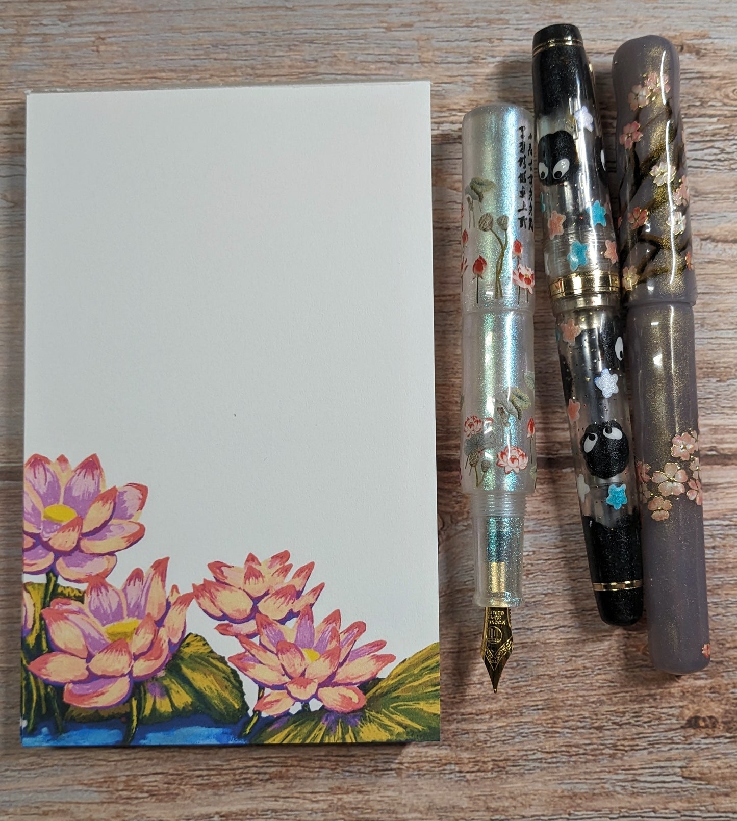 Three decorative fountain pens on a wooden surface with a notebook featuring a floral design.